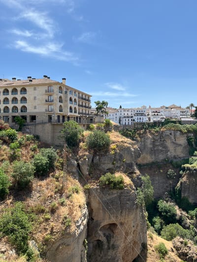 The image shows the Puente Nuevo bridge in Ronda, Spain, a historic stone bridge that spans the El Tajo gorge. The bridge is a popular tourist attraction and offers stunning views of the surrounding landscape. The bridge is made of stone and is about 120 meters long and 30 meters high. It was built in the 18th century and is a symbol of Ronda. The image also shows the city of Ronda, which is built on a hilltop overlooking the gorge. The city is home to a number of historic buildings, including the Alcazaba, a Moorish fortress, and the Plaza de Toros, a bullring. The image is taken from a high vantage point, looking down on the gorge and the bridge. The sky is blue and there are a few white clouds. The sun is shining and the weather is warm. The image is a beautiful and peaceful scene.