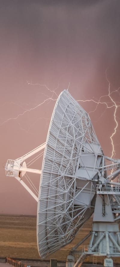 The image shows a large, white, parabolic satellite dish with a complex lattice structure. The dish is positioned at an angle, with its reflective surface facing towards the left of the frame. Behind the satellite dish, the sky is a dusky pinkish-brown, with numerous white, branching lightning bolts illuminating the scene. The lightning appears to be a significant storm in progress. In the foreground, the base of the satellite dish is supported by a sturdy, white metal structure, and the ground beneath it is a dry, tan-colored plain or desert. The overall scene evokes a sense of power and drama, with the artificial structure of the satellite dish contrasting with the natural phenomenon of the lightning storm. The image does not provide any visual cues to suggest a location in Hässelby, Sweden.