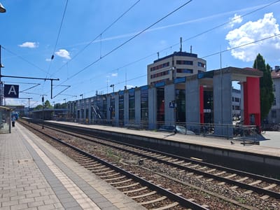 The image captures a view of the Memmingen train station in Germany on a clear, sunny day. The foreground features two sets of parallel train tracks, with ballast and some vegetation growing between them. To the left, a paved platform with tactile paving is visible, leading towards a glass-covered waiting area. A person in a blue shirt and dark pants is walking away from the camera on this platform.
In the middle ground, a modern train station building stretches horizontally. This structure has a concrete facade with large glass windows and red structural elements. Overhead, a complex network of catenary wires for electric trains crisscrosses the sky, supported by poles. A sign with the letter "A" is visible on the platform building.
In the background, a taller, multi-story building with a light-colored facade and orange-accented windows stands behind the station building. The sky is bright blue with scattered white clouds. To the right of the station building, a row of parked bicycles and a dark car are visible, suggesting an area for commuters. The overall scene depicts a functional and clean urban transportation hub.