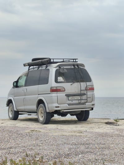 A silver Mitsubishi Delica van, equipped with a roof rack carrying a spare tire and an awning, is parked on a rocky shore next to the Caspian Sea in Aqshuqyr, Kazakhstan. The van has off-road tires and a ladder mounted on the rear. The license plate is visible and reads "KZ 686PAA 12". The sky is overcast. The environment is natural, with the sea in the background and a gravelly beach in the foreground. There are no people visible in the image.