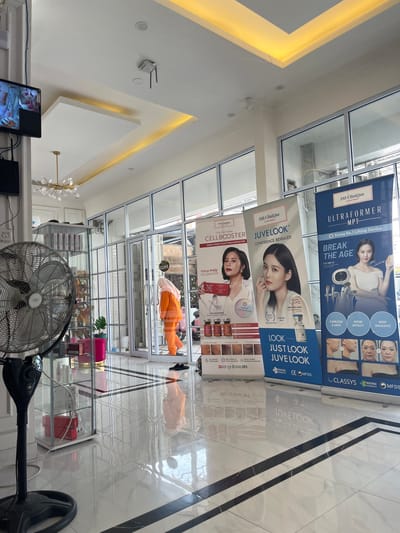The image depicts the interior of a modern clinic, likely a beauty or medical spa, located in Dumai City, Indonesia. The clinic features large glass windows and doors, a polished white tiled floor with black marble inlay, and a sophisticated ceiling design with recessed yellow lighting. Three large banner advertisements are prominently displayed in the foreground, promoting aesthetic treatments.
On the left, a large industrial-style fan is positioned next to a glass display cabinet containing skincare products. In the background, a woman wearing an orange uniform and a headscarf is seen walking through the glass doors, suggesting the clinic is either open or closing. The overall ambiance is clean, bright, and professional. The banners, particularly the one on the right, advertise a device called "ULTRAFORMER MPT" and claim it is "Korea No.1 Lifting Device," with text highlighting benefits like "PAINLESS & SAFE," "WOW EFFECT," and "NON INVASIVE." Another banner promotes "JUVE LOOK" with the tagline "CONFIDENCE REVEALED," and a third advertises "CELLBOOSTER." The presence of these specific advertisements indicates the clinic specializes in cosmetic and dermatological treatments.