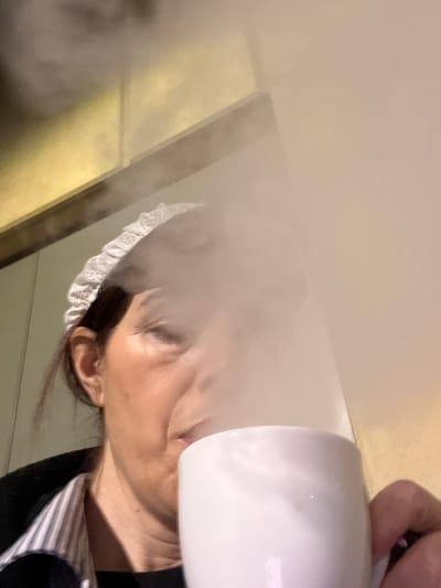 The image displays a close-up, low-angle view of an individual indoors in Roodt-sur-Syre, Luxembourg. An adult female, positioned in the lower-left frame, is seen looking upwards towards the camera. She possesses dark hair, partially covered by a white, lace-edged headband or cap. Her right eye appears partially closed or squinted. She is wearing a dark outer garment over a striped undershirt. A white ceramic mug is held in her right hand and positioned near her mouth, emitting a dense plume of white steam that obscures the central and upper-right portions of her face and the image frame. The background consists of interior wall elements, including a reflective golden panel in the upper-left, above a lighter-toned vertical surface.