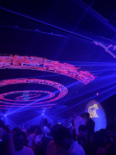 The image captures an indoor scene, likely a nightclub or bar, in Tuxtla Gutiérrez, Mexico. The dominant feature is a large video screen displaying red and orange patterns of concentric circles and abstract designs against a deep blue backdrop. Blue laser beams crisscross the space, adding to the visual intensity. A group of people, most with their backs to the camera, stands to the right of the screen, watching the display. Some are wearing hats, and a few are in light-colored shirts. Above the crowd, a round, lit sign is visible, featuring a neon or backlit design. The overall atmosphere suggests a lively, entertainment-focused environment with a strong emphasis on visual effects.