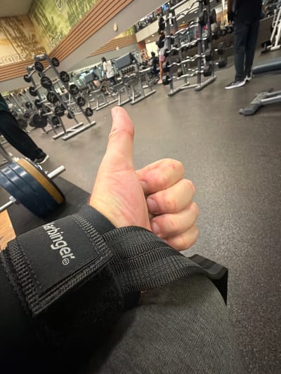 The image shows a first-person view inside a gym in Duluth, United States of America. The subject is wearing a weightlifting wrist wrap labeled "Harbinger" on their right wrist. The person's hand is visible, with a thumb extended. In the background, there are weight racks with dumbbells, and other gym equipment can be seen. The floor is gray, and the lighting appears to be artificial, typical of indoor gym environments.