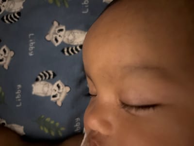 This is a close-up, eye-level, medium shot photograph of a young child's face sleeping on a blue fabric with a pattern of cartoon raccoons and the word "Libby". The child's face, from the nose upwards, fills the right side of the frame. Their eyes are closed, and their eyelashes are visible. The child's skin tone is warm and smooth. The fabric in the background is slightly out of focus, but the raccoon illustrations and the text are discernible. The scene is indoors, with soft lighting. The image is a personal photograph, likely taken by a caregiver. The location in Geulanggang Teungoh, Indonesia is not indicated by any visual cues in the image itself.