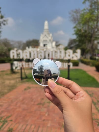 A person holds a white, circular mirror with bear ears in front of a white Thai temple and large Thai script sign in the Historic City of Ayutthaya, Thailand. The mirror reflects a person with a cell phone taking the photo. The foreground is a red brick pathway and green grass. The weather appears sunny with a few clouds.