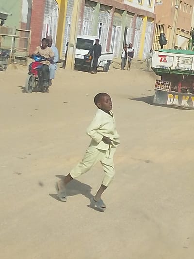 The image depicts a street scene in Mariri, Nigeria. In the immediate foreground, a young Black male child, approximately 5-7 years old, is shown mid-stride, moving from left to right across a light-colored, unpaved or dusty road. He wears a light yellowish-tan long-sleeved tunic and matching cropped pants, with light grey slip-on sandals. His gaze is directed forward and slightly upwards, with his mouth slightly ajar.
In the mid-ground to the left, two Black adult males are riding a red and black motorcycle away from the viewer. The driver wears a light brown shirt and dark pants, while the passenger behind him is clad in a light blue garment and a light-colored cap. Further left, a white pickup truck is parked, and a dark-clothed figure stands near its rear.
In the mid-ground to the right, a large truck with a green cargo bed and a red undercarriage is partially visible, displaying text including "HORN BEFORE" and "DA KYA". Several indistinct figures are visible walking on the far side of the street.
The background consists of multi-story buildings with brick and painted facades, featuring numerous windows, some with white frames and security bars. Overhead utility lines and poles are visible. The scene is illuminated by bright daylight, casting visible shadows.