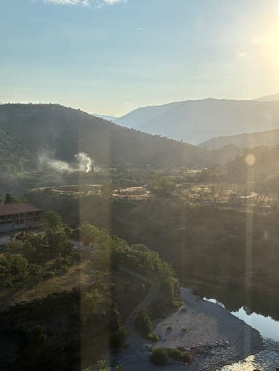 The image depicts a serene valley landscape under a bright, hazy sky, likely during sunrise or sunset, casting a warm glow over the scene. In the foreground, a wide, rocky riverbed slopes down to a calm body of water. Lush green trees and vegetation line the riverbanks and extend up the slopes of rolling hills that dominate the mid-ground and background.
Nestled within the valley, a cluster of buildings is visible. On the left, a two-story building with a red-tiled roof stands prominently. Nearby, what appears to be construction or agricultural machinery, including a yellow vehicle and a red vehicle, are present. Further into the valley, a thin plume of white smoke rises from the trees, suggesting a controlled burn or a small fire.
The background consists of layers of undulating hills and mountains, fading into a soft haze due to atmospheric conditions and the direction of the sunlight. The overall impression is one of rural tranquility and natural beauty. The location is identified as Donje Mrke, Montenegro.