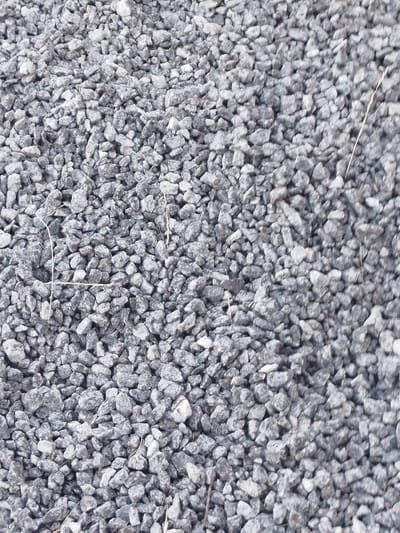 The image displays a close-up, top-down view of a surface covered in small, irregularly shaped gray stones, commonly referred to as gravel. The stones vary in size, with most being between 1 to 3 centimeters in diameter. Some lighter-colored stones are interspersed with the predominantly darker gray ones. A few thin, dried grass stalks are visible lying on top of the gravel. The image appears to be taken outdoors, with natural lighting conditions. There is no discernible information to place this scene in the city of Holendry, Ukraine. The scene is devoid of people, animals, or any discernible structures. The focus is entirely on the texture and composition of the gravel.