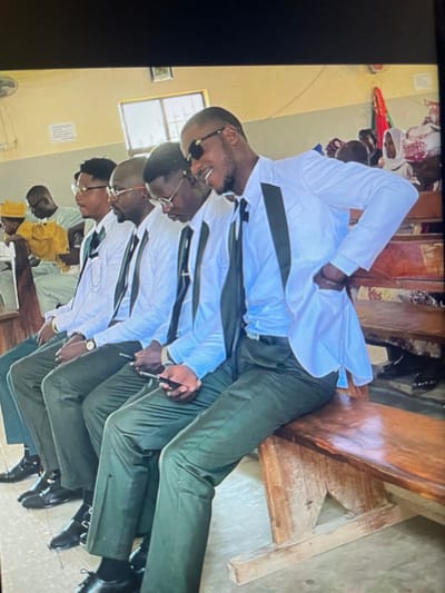 The image depicts an indoor gathering in a public hall or community center located in Mara, Nigeria. Five men, uniformly dressed in white long-sleeve shirts, dark ties, dark green trousers, and dark blazers with dark lapels, are seated in a row on a wooden bench in the foreground, facing right. The man on the far right wears sunglasses, exhibits a smiling expression, and has his right hand in his jacket pocket. The two men immediately to his left are observed looking down at smartphones held in their hands. The fourth man from the right wears eyeglasses and a wristwatch on his left wrist. The fifth man in the row, on the far left, is largely obscured. In the background, numerous individuals, including men and women, are seated on additional wooden benches. Specific attire includes light green garments, a yellow hat, a white head covering, and a colorful scarf among the background figures. The environment features light-colored walls, a multi-pane window, a partially visible ceiling fan, a framed object on a wall, and a red and green banner in the distant background. The floor appears to be concrete or tiled.