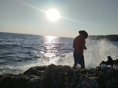 A person wearing a red shirt, dark shorts, and a dark hat stands on a rocky shoreline, silhouetted against the bright sun. The sun is high in the sky, creating a strong lens flare. Waves crash against the rocks, sending spray into the air. The water is a deep blue, and the sky is a pale blue. In the distance, there is a hazy coastline with hills. The scene appears to be in Karaburun, Turkey, likely during the day.