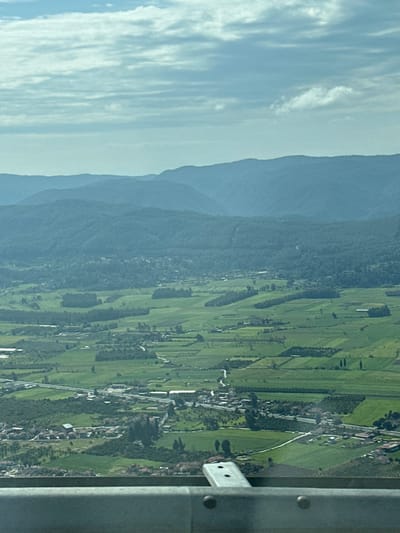 The image presents a panoramic, high-angle view of a rural landscape within Ula, Turkey. The foreground displays a metallic gray barrier or railing, secured by two visible rivets, with a perpendicular metallic bracket extending from its upper edge. The midground is dominated by a vast expanse of segmented green agricultural fields, interspersed with scattered small settlements comprised of residential and commercial structures. Multiple winding roads traverse the landscape, including a more prominent thoroughfare with buildings lining its path. Patches of cultivated land, some appearing as orchards, are visible. In the background, multiple undulating mountain ranges, covered in dense vegetation, recede into the distance, progressively lighter in hue due to atmospheric perspective. The sky occupies the upper portion of the frame, filled with a stratum of light blue-gray stratocumulus clouds. No human subjects, animals, or specific actions are discernible within the scene.