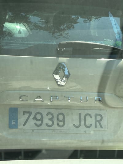 The image shows the rear of a white Renault Captur. The car has a license plate that reads "7939 JCR". The license plate is from Spain, indicated by the blue European Union band on the left with the letter 'E' for España. The car is parked outdoors, as evidenced by the reflections of trees and sky on the rear window and the bright sunlight. The location is identified as Benimàmet-beniferri, Spain. There are no people visible in the image. The scene is static and forensic in nature, focusing on the vehicle and its identifying marks.