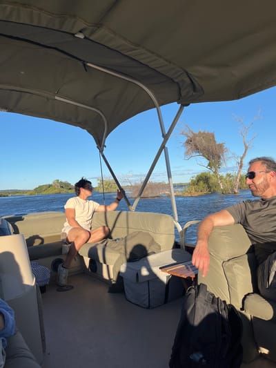 The image depicts two individuals on a boat, likely on the Zambezi River near Livingstone, Zambia, based on the environment and the presence of riverine vegetation. One man, wearing a dark shirt and sunglasses, is seated and appears to be interacting with an object on a small wooden table. The other person, wearing a light-colored shirt, seems to be reaching upwards, possibly adjusting or inspecting the boat's canopy support. The boat is equipped with upholstered seating and a canopy, suggesting a leisure activity. The river flows in the background, flanked by trees and vegetation typical of the Zambezi River ecosystem. The sky is clear and blue.