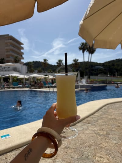 The image captures a close-up of a hand holding a light yellow drink with a black straw in a tropical resort setting. The drink is in a clear stemmed glass and appears to be a refreshing beverage, possibly a cocktail. The hand is adorned with two bracelets: one is a light, almost ivory color, and the other is a darker, amber-toned resin.

The background reveals a vibrant blue swimming pool, hinting at a resort or hotel environment. Several lounge chairs with white canopies are visible along the poolside, suggesting a place for relaxation. In the distance, there are palm trees and a hillside covered in greenery, characteristic of a warm, sunny location. The sky is a clear blue with scattered white clouds, indicating fair weather. The overall scene evokes a sense of vacation and leisure.

A tattoo of the numbers "1986" is visible on the forearm holding the drink. The presence of a building with multiple levels and balconies in the background further reinforces the idea of a resort or hotel. The image is oriented vertically, with the drink and hand in the foreground and the pool and landscape extending into the background. The lighting suggests it is daytime, likely late morning or afternoon, given the bright sunlight.