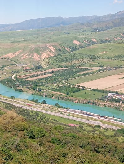 The image presents a landscape in Chinarkent, Uzbekistan. The scene features a river of turquoise water flowing alongside a highway. The highway has multiple lanes and shows some vehicle traffic, including a red truck. Adjacent to the highway is a fenced area with industrial structures.
The landscape includes green hills and mountains in the background, with some exposed earth areas visible on the hillsides. The vegetation in the foreground consists of shrubs and trees. The terrain appears to be varied, with agricultural fields and some buildings interspersed among the natural landscape. The sky is clear and blue.
