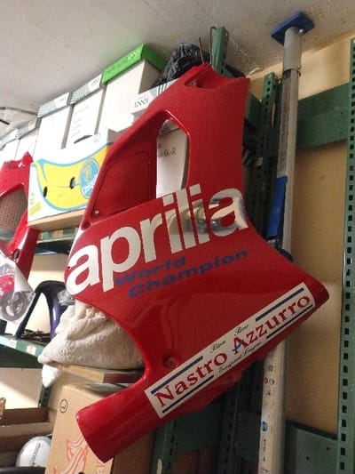 The image displays a storage area within a building in Fentange, Luxembourg. The central focus is a glossy red motorcycle fairing, likely a side panel, angled towards the viewer. It features prominent white "aprilia" lettering, "World Champion" in blue text below, and a rectangular sticker displaying "Nastro Azzurro" in bold text, with "Birra Beer" written above it.
In the background to the left, multiple cardboard boxes are stacked on shelves; one box is branded "Chiquita". Other red motorcycle components are partially visible. A light-colored, crumpled material rests behind the fairing. Further down, a white spherical object with an indistinct dark logo is partially visible.
To the right, light green metal shelving units are visible. A silver-colored adjustable support pole with a blue top, marked "SAM", extends vertically from the floor to the ceiling, adjacent to the shelving. The wall is light beige, and the ceiling is white.
No individuals are present, and no actions or interactions are depicted.