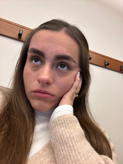 The image is a close-up, portrait-style photograph of a young woman, taken from a slightly upward angle and rotated counter-clockwise. The woman's head and upper torso dominate the frame.

She has long, straight brown hair, parted in the middle, which cascades down her shoulders and across the lower part of the image. Her complexion is fair, and she has prominent, well-defined dark eyebrows. Her eyes are brown and appear to be looking directly or slightly to the side of the camera, with a neutral or contemplative expression. Her lips are a natural pink hue, slightly parted.

Her right hand (from the viewer's perspective) is raised, with her fingers gently resting against her cheek and jawline. She is wearing white nail polish on her visible fingernails. A silver-toned ring with a clear or light-colored stone is visible on her right ring finger. She is dressed in a light-colored, possibly beige or light pink, knitted sweater with a textured, ribbed pattern. A white long-sleeved top or undershirt is visible beneath the sweater at the wrist cuff.

The background is a plain, light-colored wall, likely white or off-white. A narrow, dark wooden panel or strip runs diagonally across the upper left portion of the background. Attached to this wooden strip are several dark-colored hooks or clips, evenly spaced. These hooks appear to be coat hooks or similar utility fixtures. The lighting is bright and even, suggesting an indoor setting. There is no visible text in the image.