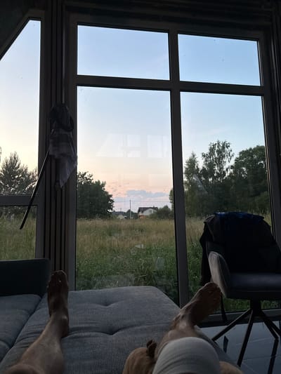 This image depicts a person's legs resting on a sofa, looking out of a large window. The window frames a rural landscape with tall grass, trees, and houses in the distance, suggesting the location is the city of Vol'noe, Russia. The sky is a pale blue with hints of pink and orange, indicating either sunrise or sunset. A chair with clothing draped over it is visible to the right of the frame. A patterned cloth and a belt hang from the window frame on the left. The overall scene conveys a sense of relaxation and domesticity.