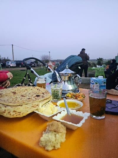 An outdoor table is laden with food. Flatbreads, butter, honey, olives, a dessert, and a glass of tea are arranged on a tan surface. A silver teapot and a glass with a football pattern are nearby. A stroller and people are visible in the background, suggesting an outdoor gathering. The sky is overcast.