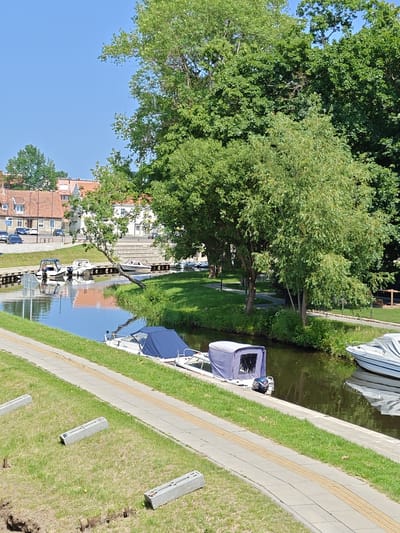 The image depicts a tranquil canal scene in Šilutė, Lithuania, on a clear, sunny day. Several boats are moored along a wooden jetty on the left side of the canal. These include a white motorboat with a blue cover, a smaller white boat with a dark blue canopy, and another white boat with a blue canopy further down the canal. On the right side of the canal, a white boat covered with a grey tarp is visible.
The canal is lined with lush green grass and a paved walkway with tactile paving. In the background, a row of buildings with red-tiled roofs and white facades suggests a residential or commercial area. Mature trees with dense green foliage line the canal banks, casting shadows on the water. The water is calm, reflecting the blue sky and the surrounding greenery. The scene conveys a sense of peacefulness and outdoor recreation. No people are visible in the image.