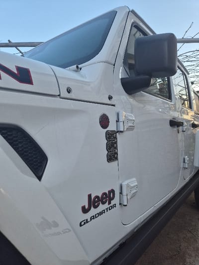 The image captures the front passenger side of a white Jeep Gladiator pickup truck, viewed from a low angle. The vehicle exhibits a red and black "Jeep" emblem with "GLADIATOR" lettering directly below it on the front fender. A dark ventilation grille is present on the hood, which also bears partial red "RUBICON" lettering. A circular red badge and multiple stacked black rectangular badges are affixed to the fender. The vehicle features a black side mirror, exposed silver-colored door hinges, and a black running board along its lower edge. The windshield reflects a clear blue sky. In the background, a metallic pole structure and bare tree branches are visible. No individuals are present, and no actions or interactions are depicted. The image contains no discernible visual markers confirming a specific location within Balsha, Bulgaria.