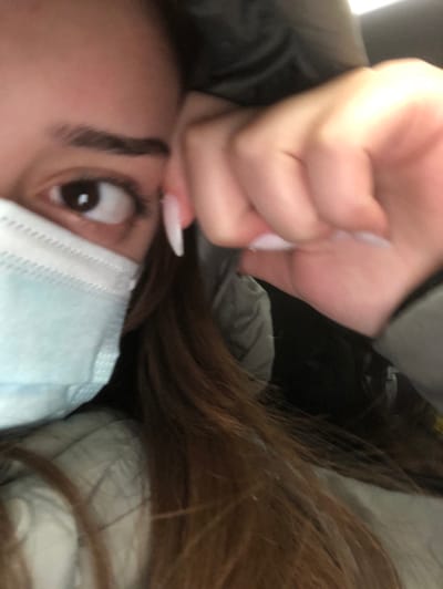 A close-up, rotated image depicts a person's upper body and face from an unusual, oblique perspective. The subject's right eye, wide open and brown, looks directly towards the camera, suggesting surprise or intensity. A well-defined eyebrow is visible above the eye. The person is wearing a light blue surgical mask, covering their nose and mouth.

Long, dark brown hair cascades across the right side of the frame. In the upper portion of the image, a hand is clenched into a fist, positioned near the person's head. This hand features long, pointed fingernails with a light, possibly white or pale pink, polish. The person appears to be wearing a dark jacket or coat, with its material visible on the left.

The background is dark and indistinct, hinting at an indoor setting or a confined space, possibly a vehicle interior. The composition suggests a selfie taken from a low and close angle. No specific time of day or weather conditions can be determined from the image. While the provided context indicates Steinbourg, France, there are no visual cues within the image itself to confirm this location. No text is visible.