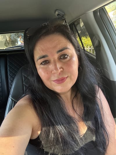 The image shows a woman sitting in the driver's seat of a car. She has long, dark hair and is wearing a black top. Her face is partially visible, and she is looking towards the camera with a slight smile. Sunglasses are perched on top of her head. The interior of the car is visible, including the black leather seat and part of the dashboard. Through the rear window, two people can be seen in the backseat, with one appearing to be a child. Outside the car, trees and a fence are visible, suggesting a suburban or natural environment. The lighting suggests it is daytime, possibly late afternoon due to the warm sunlight. The specific location in Novotroitsk, Russia, cannot be determined from the visual information provided in the image.