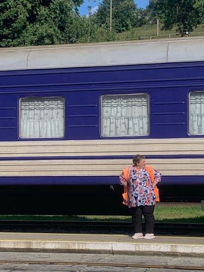 A forensic description of the scene depicts a middle-aged woman standing on a concrete train platform at the Yasinia train station in Yasinia, Ukraine. The woman, with graying hair, wears a multi-colored floral tunic, black trousers, light-colored sneakers, and an unzipped orange high-visibility vest. Her hands are placed on her hips as she looks towards the right side of the frame. Behind her, a dark blue passenger train car with light-colored horizontal stripes and multiple windows fitted with patterned curtains is parked on the tracks. The train roof is light grey. In the background, a hillside covered with dense green trees rises, with a clear blue sky visible above. Below the platform, train tracks are discernible. The scene is illuminated by natural daylight.