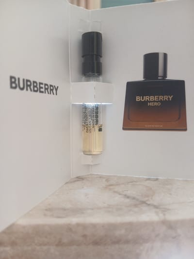 This media file is a close-up shot of a Burberry Hero fragrance sample. The sample is presented in a white cardboard sleeve, with the Burberry logo prominently displayed in large, gray lettering on the lower left side. Above this, there's an image of the full-size Burberry Hero fragrance bottle, rendered in shades of brown and black, with the text "BURBERRY HERO" and "ELIXIR DE PARFUM" visible. To the right of this image, a small, clear vial containing a golden liquid is held in place by the cardboard. The vial has text printed on it, which appears to be product information and distribution details, including "MADE IN FRANCE" and "US/CAN: 1-800-715". The background is a neutral, textured surface, possibly a countertop or table, with subtle variations in color and pattern, suggesting an indoor setting. The lighting is even, indicating it was taken in a well-lit environment, likely indoors, with no specific time of day or weather conditions discernible. The overall presentation is neat and organized, focusing on the product itself.