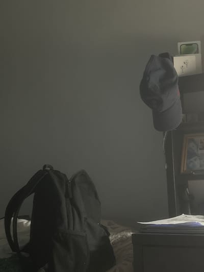 The image depicts a dimly lit bedroom interior. In the foreground on the left is a black backpack, open and resting on a bed. To the right, a dark-colored baseball cap hangs from a hook or string, suspended in mid-air. Behind the cap, a dark shelf unit is visible. On the top shelf of this unit are two white boxes, one with a green image suggesting electronics, and another with an image of white earbuds. Below these, a framed photograph or picture hangs on the shelf unit, showing what appear to be faces. Further to the right and slightly lower, a portion of a dark wooden dresser or table is visible, with some papers or documents lying on top. The overall lighting is low, casting shadows and obscuring some details. The environment appears to be a private residential space, possibly a bedroom, in Carrières-sous-Poissy, France.