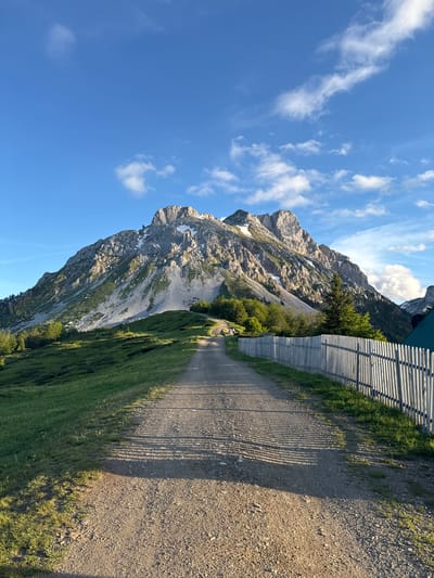 The image depicts a scenic mountain landscape with a dirt road leading towards a rugged, rocky mountain peak. The sky is a clear blue with scattered white clouds. On the right side of the road, a white wooden fence runs along the grassy slope. The mountain is partially covered in snow, and there are patches of green vegetation on its lower slopes. The road is well-trodden and casts long shadows from the fence. In the distance, several trees are visible on the left side of the road. This scene is located in Jošanica, Montenegro, a region known for its natural beauty and mountainous terrain.