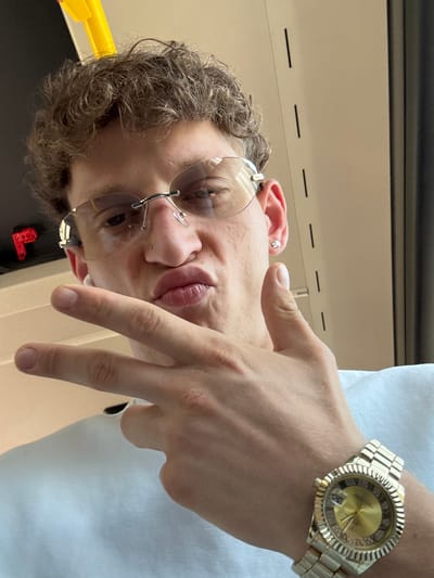 The image shows a close-up, vertically oriented shot of a young person's face and hand. The subject, facing upwards and slightly to the left, has curly brown hair, and is wearing clear, rimless glasses with silver frames, and an earring in their left ear. A nose piercing is also visible. Their lips are parted, and they appear to be making a peace sign with their right hand, which is in the foreground. On their left wrist, they are wearing a gold-colored watch with a fluted bezel and a diamond-studded face. The watch face has Roman numerals and a date window. The background appears to be indoors, with a neutral-colored wall or surface and some horizontal lines. A yellow object is partially visible in the lower left corner. The overall lighting suggests it is daytime. There is no visible text in the image.