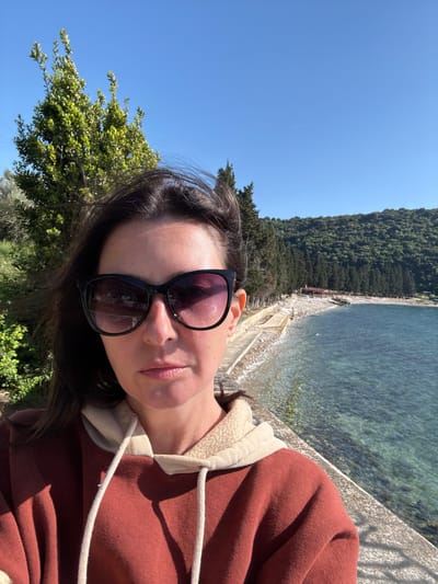A woman takes a selfie outdoors in Bratica-Braticë, Montenegro. She wears a burgundy hoodie and sunglasses. Behind her, a concrete barrier overlooks a beach and turquoise water. A dense forest covers the hillside along the coastline. The sky is clear and blue. Green foliage grows in the foreground.