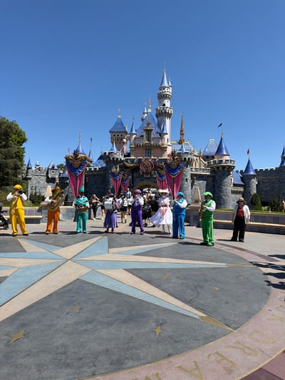 This image captures a daytime scene at Disneyland Park in Anaheim, California, with Sleeping Beauty Castle serving as the prominent backdrop. In the foreground, a group of seven performers dressed in colorful, coordinated costumes are arranged on a large, star-shaped pavement design. The performers are playing various brass and percussion instruments, suggesting a marching band or musical ensemble.
From left to right, the performers are:
1. A person in a bright yellow vest and matching pants, playing a trumpet.
2. A person in an orange vest and matching pants, holding a tuba.
3. A person in a teal vest and matching pants, playing a drum.
4. A person in a purple dress and white hat, holding a cymbal.
5. A person in a purple vest and matching pants, playing a clarinet.
6. A person in a green vest and matching pants, playing a saxophone.
7. A person in a white shirt, red polka-dot vest, and black pants, wearing a straw hat and observing the scene.
Behind the performers, the iconic gray stone structure of Sleeping Beauty Castle is visible, adorned with blue turrets and golden spires. Banners with "70" in large numerals and Mickey Mouse silhouettes are hanging from the castle. The sky is a clear, bright blue. Several other park visitors are visible in the middle ground, walking and standing, some blurred due to motion. The overall atmosphere suggests a festive event or performance taking place in the park.