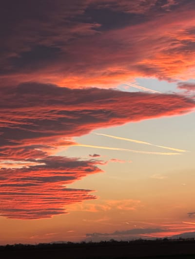 The image shows a low-angle, daytime view of red-tinged, vertical cloud formations resembling mountain peaks in the sky. Contrails are visible in the open sky above the clouds. The lower right of the image shows a blurry, dark edge. There is no evidence of Malenovo, Bulgaria. There are no people or subjects.