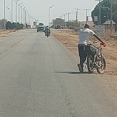 The image captures a daylight scene on an asphalt road in the city of Haifam, Nigeria. In the right foreground, an adult male, wearing a white shirt, dark trousers, and a dark head covering, pushes a dark motorcycle forward on the road's shoulder, grasping the left handlebar. The motorcycle features red handlebar accents and a dark bag secured to its rear. The subject casts a distinct shadow to his left. In the midground, another individual operates a motorcycle moving away from the viewer on the main thoroughfare. Further distant, a light-colored sedan with roof-mounted items also proceeds in the same direction. The two-lane road is bordered on the left by a linear arrangement of modern streetlights and on the right by utility poles with overhead lines. The surrounding environment consists of dry, sparse vegetation and indistinct distant structures under a hazy sky.