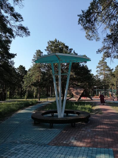 The image depicts a park scene in the city of Svirsk, Russia. In the foreground, a curved wooden bench surrounds a central structure with a teal canopy supported by white pillars. The bench is situated on a pathway paved with interlocking bricks, alternating between shades of blue-gray and reddish-brown. To the right of the bench, a playground area is visible with swings and climbing structures. An elderly person, dressed in a dark maroon coat and using a cane, walks away from the camera on the reddish-brown pathway. The background is dominated by a dense forest of tall pine trees under a clear blue sky. The lighting suggests it is daytime, likely during the morning or afternoon.