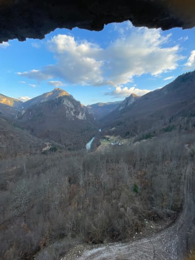 The image presents an elevated, panoramic view of the Tara River Canyon in Đurđevića Tara, Montenegro. The immediate foreground is a dark, irregular rock overhang spanning the top of the frame. Below this, a vast valley unfolds under a blue sky adorned with scattered white cumulus clouds.
Steep, rugged mountains, primarily covered with bare deciduous trees, dominate the mid-ground and background, suggesting a late autumn or winter season. A light blue-green river, the Tara River, snakes through the deepest part of the valley floor. Adjacent to the river, a small cluster of buildings with visible roofs indicates a localized settlement. Peaks in the distance feature rocky exposures. The lower right section of the image reveals a dirt road or track with visible tire impressions and scattered debris, bordering a forested incline. No individuals, vehicles, or dynamic actions are present in the scene.