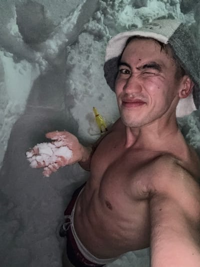 A shirtless man in a sauna hat lies on his side, holding a handful of snow. He winks at the camera with a playful expression. A small, yellow object rests on the snow near his arm. The background is filled with snow or ice, suggesting a cold environment or ice bath setting. He is wearing briefs.