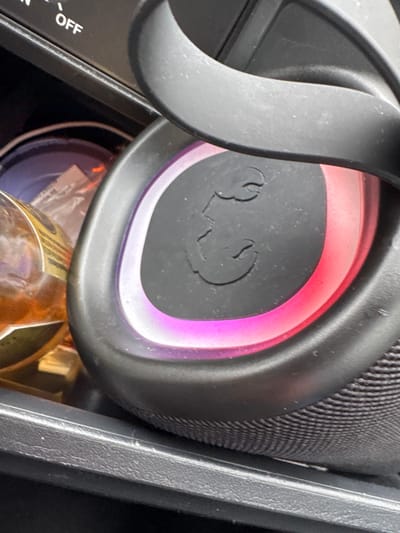 The image shows a close-up of a black portable speaker with illuminated pink and red lights on its side. The speaker has a textured surface and a raised circular logo with a stylized "E" or similar symbol. To the left of the speaker, a portion of a glass bottle with a yellow and black label is visible. In the background, a black dashboard with the word "OFF" is partially visible. The scene appears to be an interior, likely a car, given the dashboard elements and the dark, textured surroundings which could be car interior panels. The lighting suggests the speaker is actively powered on and possibly in use. The location of Marsum, Netherlands, does not directly correlate with any visible elements in the image; the provided details are purely based on the visual content of the photograph.