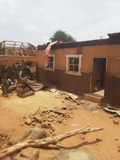 The image shows a damaged building in Kwal, Nigeria, located at coordinates 9.804 latitude and 8.647 longitude. The building appears to be a single-story, residential structure with an orange facade. The roof is partially collapsed, with debris scattered around the exterior. A large piece of corrugated metal is hanging from the roof, suggesting a strong wind or storm. Several pieces of wood are visible, possibly part of the roof structure. There are two windows and a door on the exterior of the building. The windows have glass panes, but the panes are broken. The door is open, revealing a dark interior. The surrounding ground is covered in dirt and debris, suggesting a recent event. There are several large rocks and some tree limbs scattered on the ground. There are no people or animals in the image. The image appears to have been taken in daylight. The sky is clear with little cloud cover.