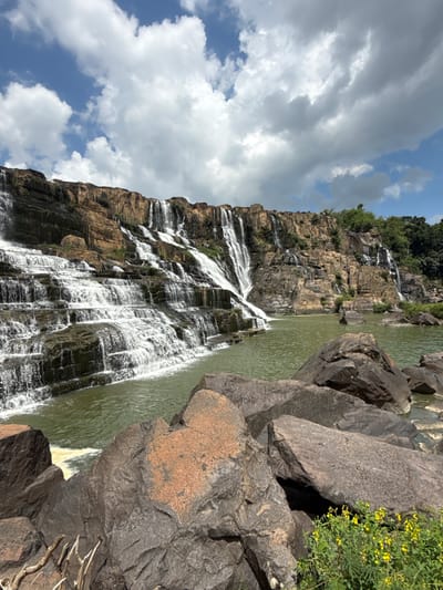 The image depicts a multi-tiered waterfall cascading down a rugged, rocky cliff face. The water is a vibrant green, collecting in a pool at the base of the falls. Large, dark rocks are scattered in the foreground, with a cluster of small yellow flowers growing among them. The sky above is a bright blue, dotted with fluffy white clouds. Lush green trees line the top of the cliff and extend to the right of the frame. The scene appears to be the Pongour Waterfall in the city of Tân Phú, Vietnam.