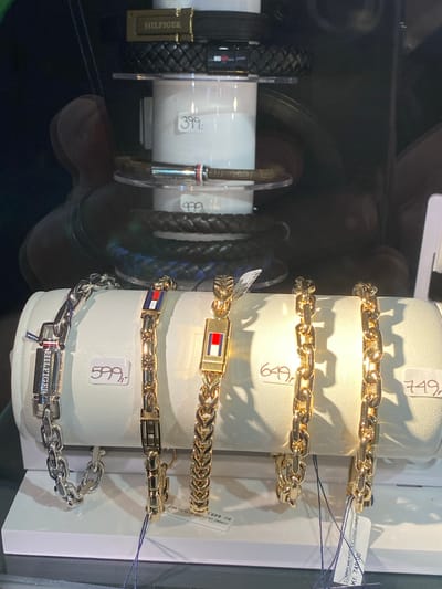 This image depicts a display case in what appears to be a retail store, likely a jewelry store or department store, showcasing various Tommy Hilfiger bracelets. The bracelets are arranged on a white, curved surface. On the left, a silver-colored chain bracelet with a "HILFIGER" inscription is visible, priced at 599. Next to it are gold-colored bracelets, one featuring small Tommy Hilfiger flags and priced at 649. Further to the right, another gold-colored bracelet is displayed, with a price tag of 749. In the background, higher up in the display, a black braided belt with a gold "HILFIGER" buckle is visible, along with other braided bracelets and some pricing tags showing 399 and 499. Reflections of people are present in the glass of the display case, suggesting a public space. The context suggests this is a retail environment where these accessories are for sale. Given the mention of Herning, Denmark, this scene is likely from a store within that city.