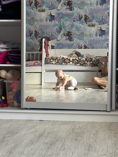 The image shows a reflection in a mirrored closet door. A baby is crawling on the floor in the reflection, wearing a yellow shirt and white pants. The baby is playing with a toy box. To the right of the baby, a person's legs are visible, suggesting they are sitting on the floor. In the background of the reflection, there is a bed with a floral patterned comforter and a wall covered in wallpaper featuring characters from the movie Frozen. To the left of the baby, a toy bin filled with various toys is visible, as well as a stuffed animal. The closet itself contains stacked clothing and bags. The setting appears to be a child's bedroom. There is no visual information in the image that can confirm or deny the location of Мариинский, Russia.