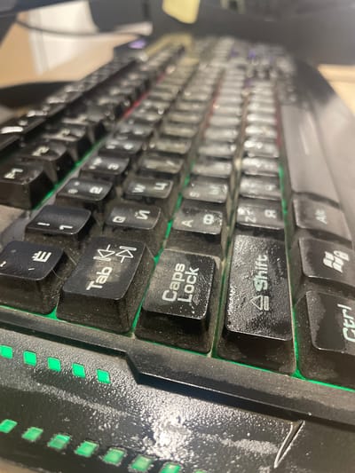 The image shows a close-up, low-angle view of a dirty, black computer keyboard with green backlighting. Several keys are visible, including "Tab," "Caps Lock," "Shift," and "Ctrl." There are also keys with what appear to be Cyrillic characters and numbers. Dust is visible on the keys and the surrounding keyboard surface. The lighting suggests an indoor environment, possibly an office or home setting. There are no people or discernable city-related features present in the image. The location of Терволово, Russia, is not visually represented in this photograph.