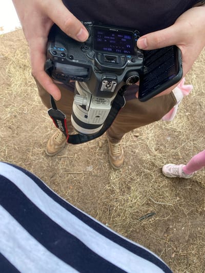 The image shows a close-up view of a person holding a Canon camera and a smartphone. The camera has a Canon brand lens attached. The person is wearing brown pants and tan shoes. A small child's pink leggings and sneakers are visible in the lower right corner of the image. The background appears to be an outdoor environment with dry grass and dirt. The image does not contain any information that would allow for its localization to Baltiiskoe, Russia.