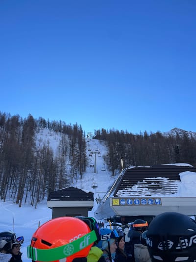 The image captures a bright, clear day at a bustling ski resort, likely Courmayeur, Italy. The scene is dominated by a brilliant, cloudless blue sky filling the upper portion, suggesting excellent weather conditions for skiing.

In the foreground, several individuals are visible, likely skiers or snowboarders, waiting at what appears to be a ski lift station. Their heads and shoulders are shown, equipped with helmets and goggles. A prominent feature is a person wearing a vibrant orange helmet with a lime green strap, displaying the brand "POC" and partially visible text "DIF" on the strap. Another individual wears a dark helmet with "SKI" visible and blue goggles.

To the right, a multi-story building, presumably part of the ski lift infrastructure, stands with a dark, corrugated roof partially covered in fresh snow. On its side, a series of informative signs are visible: several blue square panels with white pictograms illustrating permissible activities or passengers for the lift, including a person boarding, two people on a chair, a person with a dog, and a person with a bicycle. Below these, a yellow circular sign with the number "1" in black is displayed.

Beyond the building, a snowy ski slope extends into the background, flanked by numerous bare trees, their dark branches contrasting with the white snow. Further up the slope, several ski lift towers and chairs are visible, ascending the mountain. In the far background, rugged, snow-capped peaks rise against the clear sky. The overall impression is one of a lively winter sports environment on a beautiful, sunny day.