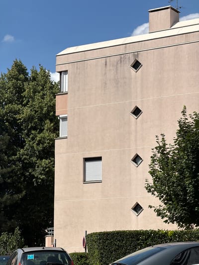The image depicts the exterior of a building, possibly an apartment complex, on a clear, sunny day. The building's facade is a light, neutral color, with vertical lines dividing sections of the wall. A notable architectural feature is the presence of several diamond-shaped windows, arranged in a diagonal pattern. One larger rectangular window with blinds is also visible.
At the top of the building, a small, box-like structure with an antenna juts out. Lush green trees partially obscure parts of the building, with their branches reaching towards the facade. To the right of the building, a dense hedge runs along the side. The lower portion of a car is visible on the far right, suggesting a street or parking area. In the lower left of the frame, a large tree fills the space, contrasting with the clear blue sky.
The lighting suggests it is daytime, and the shadows cast by the trees and the car indicate a bright, sunny atmosphere. The scene appears to be in a residential or urban setting. No people are visible in the image. There is a small, circular sign with a red border and a diagonal line through it, often indicating a prohibition, visible on the hedge. A sticker with a blue background and white symbols is on the rear window of the car. The location context of Évry-Courcouronnes, France, places this scene in a specific geographical area.