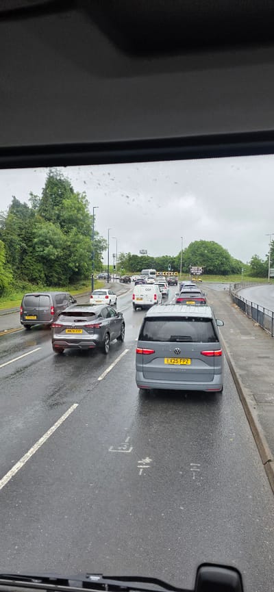 The image captures a traffic jam on a wet road in Tameside, United Kingdom. The scene is viewed from inside a vehicle, looking forward through the windshield, which is dotted with raindrops. Several cars and vans are visible on the multi-lane road, all stationary or moving very slowly.
In the foreground, a grey Volkswagen Multivan with license plate EX25 FPZ is prominent. To its left, a grey Nissan Qashqai with license plate MW24 XYP is also visible. Further to the left, a dark grey panel van with license plate TT21 HSB is in the adjacent lane. In the middle distance, more vehicles, including white vans and cars, are seen. A white van with "CGP" branding is also present. The road is wet, reflecting the overcast sky. Green trees and bushes line the left side of the road, while a metal fence and pavement are on the right. In the distance, traffic lights and signage are visible, indicating a junction or roundabout. The overall impression is of a typical congested commute on a rainy day in an urban or suburban setting.